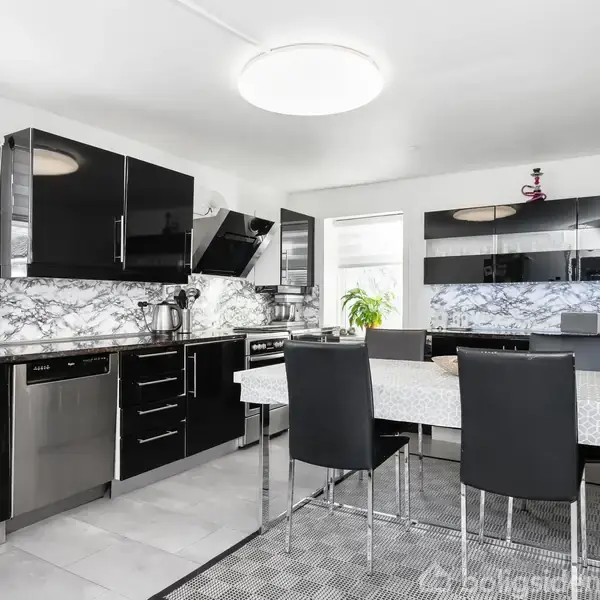 Et moderne køkken med blanke sorte skabe. Et bord med fire sorte stole står i midten. Bagved er der et marmormønstret køkkenbord og hvide vægge. En stor loftlampe lyser rummet op.