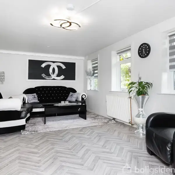 En stue med moderne møbler; en sort sofa står ved en væg, dekoreret med indrammet billede, bløde sæder og lyskilde. Flere vinduer og vægure findes i rummet.