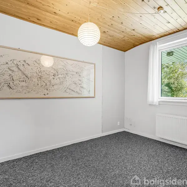 Et tomt rum med gråt tæppe, træloft og en hængende rund lampe. En stor indrammet tegning hænger på væggen. Et vindue viser træer udenfor, under en grøn tagudhæng.