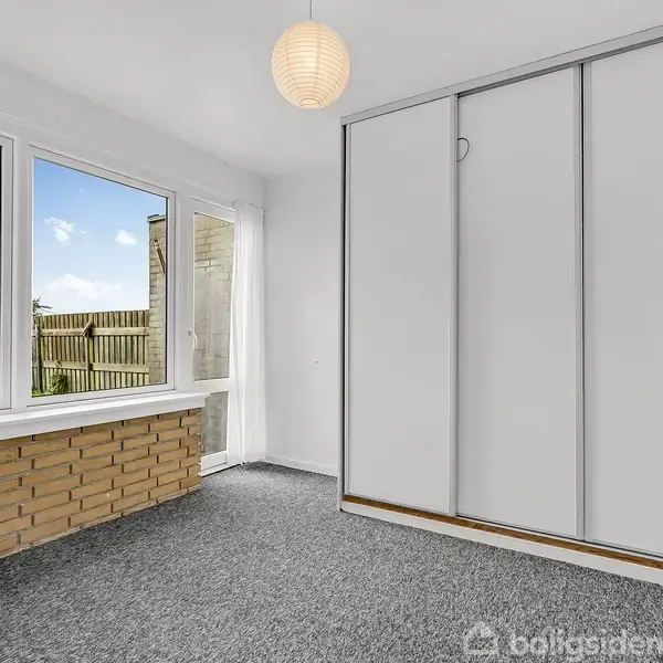 Et lyst værelse med gråt tæppe og en stor skydedørsgarderobe til højre. Store vinduer viser en have med græs og hegn udenfor. En papirlampe hænger fra loftet.