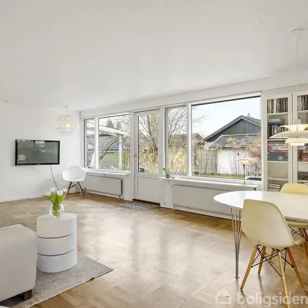 Stue med hvide vægge og trægulve. Sofa og bord er arrangeret omkring et fladskærms-tv. Spisebord med stole ved store vinduer, der viser en have udenfor.