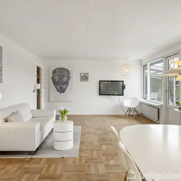 En moderne stue har en hvid sofa ved siden af et rundt sofabord med blomster; vinduerne slipper naturligt lys ind. En fladskærm hænger på væggen overfor sofaen.