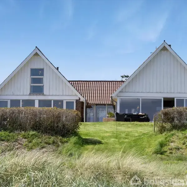 Et spisebord i haven har sort møblement med tæpper og en glasvase, placeret i nærheden af en frodig hæk. Havudsigt med bølger under en klar blå himmel i baggrunden.