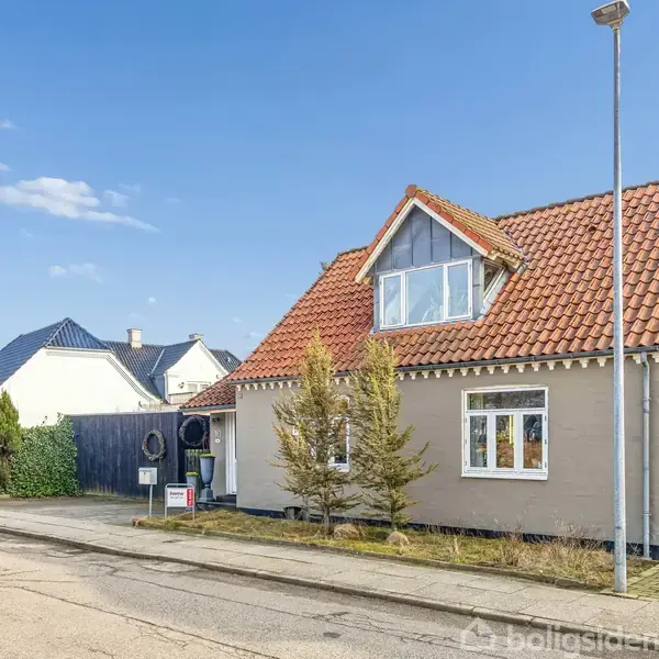 Et beige hus med rødt tegltag og stor kvist står langs en vej. To små træer dekorerer forhaven. Nabohuse og en blå himmel danner baggrunden.
