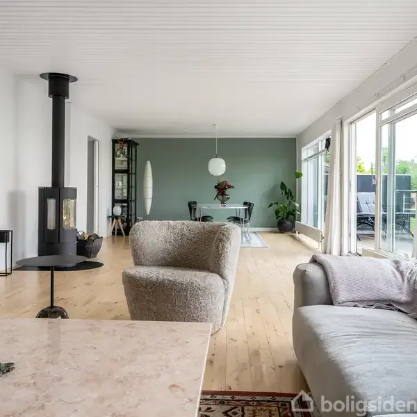 En stor glasdør fører ud til en balkon med kurvemøbler og planter. Rummet har trægulv og hvidmalede vægge. Udenfor ses grønne træer og en blå himmel.