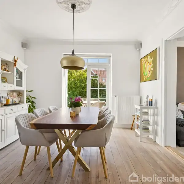 Spisebord med seks stole står i et lyst spisestue. En hængelampe over bordet, en hvid skænk til venstre og en stor vindue viser grønne buske udenfor.