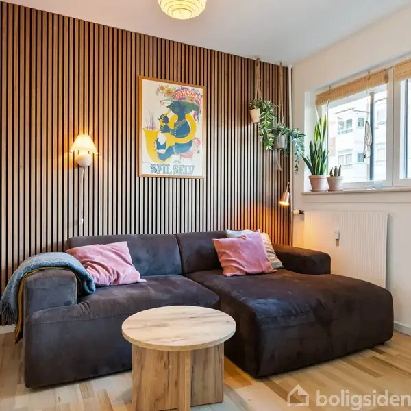 En sort sofa står mod en trævæg med en plakat med teksten "Spil Sket." Sofaen har puder og et tæppe. Til højre er et vindue med planter.