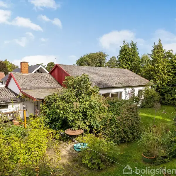 En bungalow omgivet af en frodig have med træer og buske. Bygningen har et skråt tag og røde gavle. Himlen er blå med få skyer.