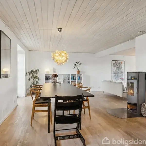 Et spisebord med stole er placeret i midten af en lys stue. En moderne brændeovn brænder i hjørnet ved siden af en plante og reol. Væggen har kunstværker.