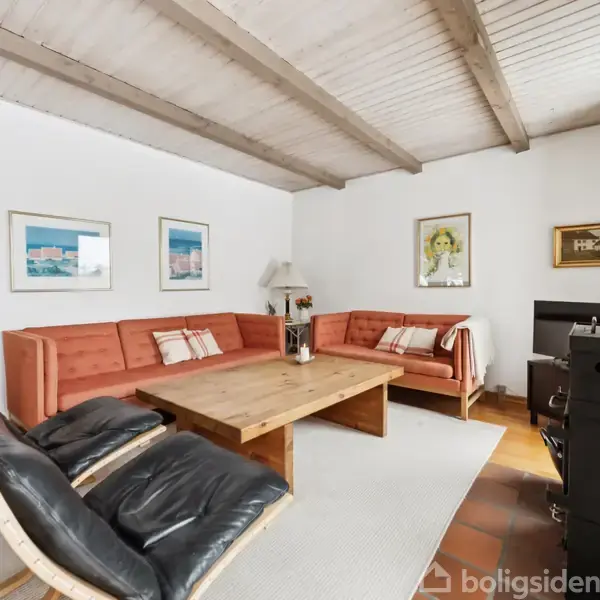 En stue med to sorte lænestole og en stor orange sofa, der omkranser et stort træbord. Rummet har hvide vægge med kunst og en sort brændeovn i hjørnet.