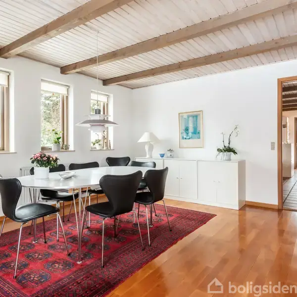 Et spisebord med sorte stole står på et rødt tæppe. Omgivelserne inkluderer lyse vægge, træbjælker i loftet og vinduer, der slipper lys ind. En hvid skænk står ved siden af bordet.