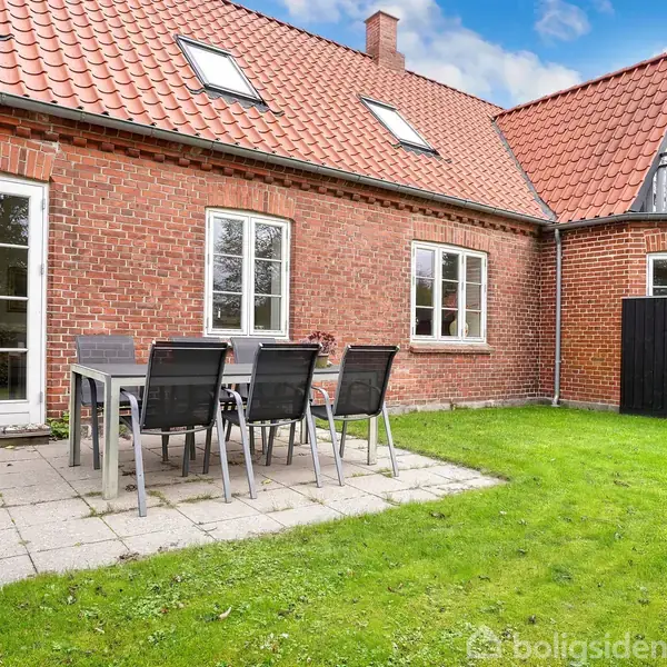 Et rødt murstenshus med rødt tegltag. En terrasse med et havebord og stole står ud for åbne franske døre. Græsplæne omgiver terrassen.