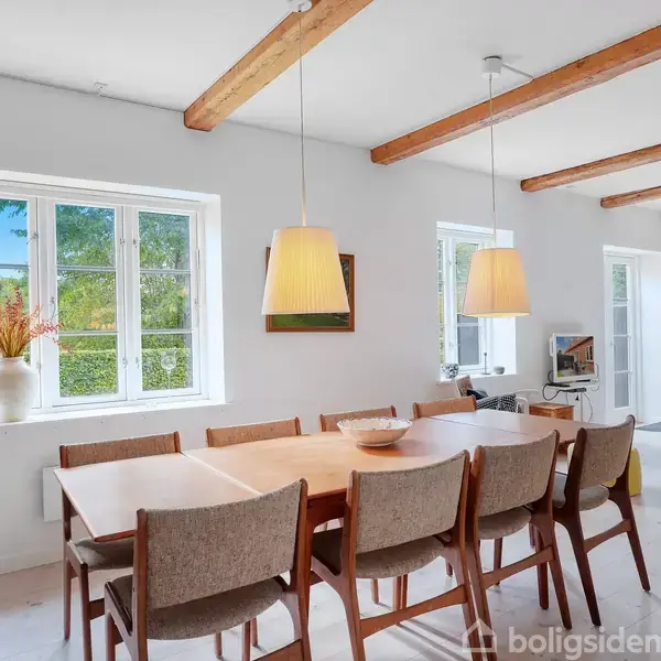 Et spisebord med otte stole står i et lyst rum med træbjælker i loftet. To hængelamper hænger over bordet. Et vindue med udsigt til en have ses i baggrunden.