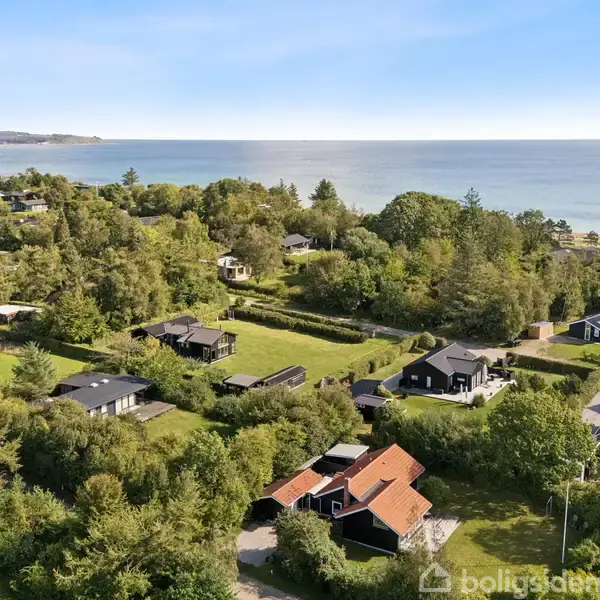 Huse ligger spredt mellem træer i et grønt område nær en kystlinje med roligt vand. Store områder med vegetation omgiver husene, der danner en lille bebyggelse.