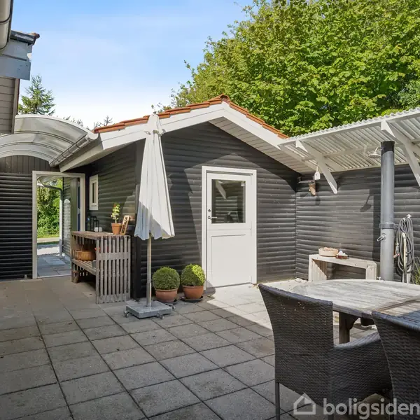 Terrasse med grå fliser har et spisebord med stole, en parasol og planter i krukker. Omgivet af sortmalede trævægge med et hvidt dørparti. Placeret i en grøn, udendørs omgivelse.