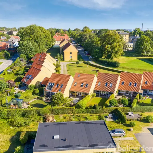 Rækkehuse med røde tage står på rad og række, omgivet af velplejede haver. En vej og grønne områder ses i baggrunden, hvor større bygninger indrammer den idylliske scene.