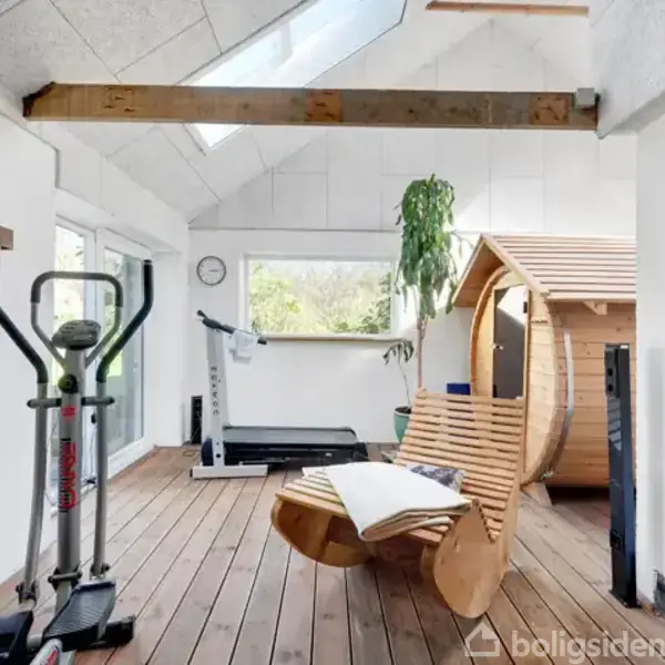 Et træningslokale med træningsmaskiner og en træsauna. En loungestol med et tæppe står foran en væg med grønne planter. Store vinduer og lyst loft giver naturligt lys.