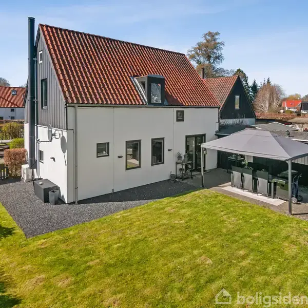 Et tofamiliershus står i en have med en grøn græsplæne. Ved siden af huset er en overdækket terrasse med sorte stole. Baggrunden viser flere huse og blå himmel.