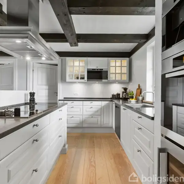 Køkkenet har hvidmalede skabe og sorte bordplader, med indbygget kogeplade. Stålelementer skaber et moderne look. Trægulv og synlige loftsbjælker giver charme. Pynt og redskaber står på bordet.