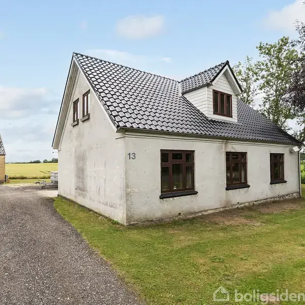 Huset er en ældre, hvid to-etagers bygning med gråt tag, placeret ved en grusvej omgivet af grønne arealer og en lysblå himmel. Nummer 13 ses på væggen.