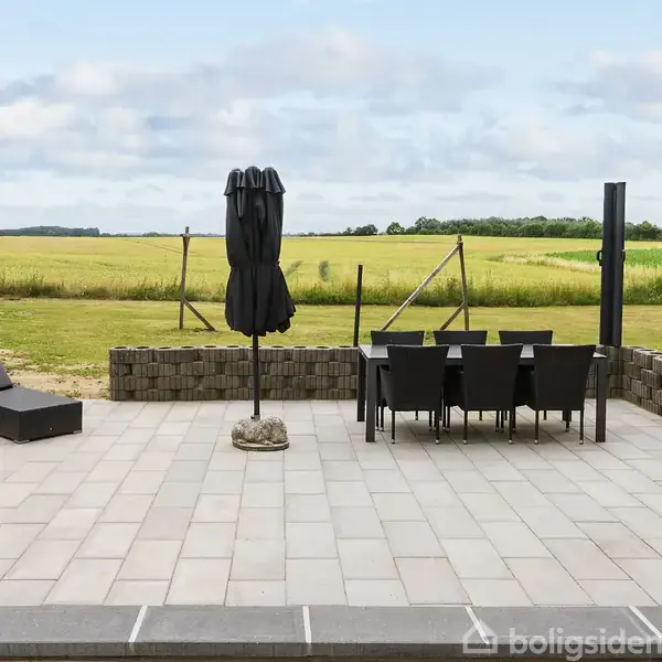 Terrasse med fliser, hvor en sort parasol står sammen med bord og stole, omgivet af lave stenvægge. En liggestol er tæt på. I baggrunden ses en mark og træer.