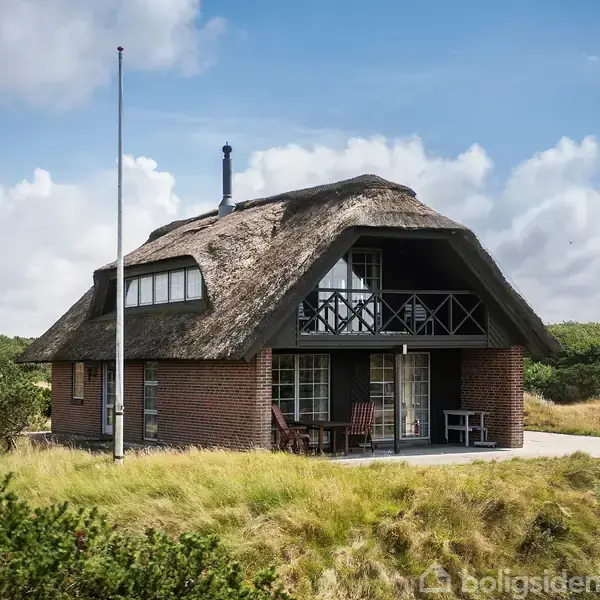 Et stråtækt hus står på en græsbeklædt grund. Der er store vinduer og en balkon. Omgivet af buske og en klar blå himmel med skyer. Flagstang foran huset.