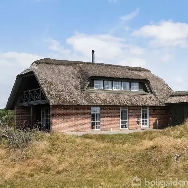 Et stråtækt hus står stille i en naturlig, græsdækket klitlignende landskab med blå himmel og få skyer i baggrunden.