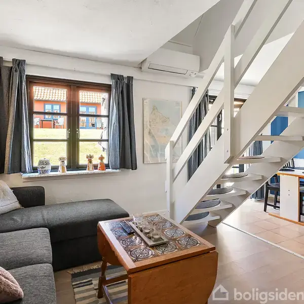 En stue med en grå sofa og sofabord. En trappe fører til en øvre etage. I baggrunden ses et spisebord med stole. Rummet er lyst med store vinduer.