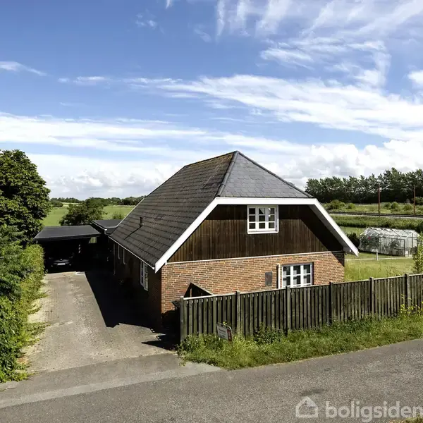 Et rødt murstenshus med et skråt tag står stille, omgivet af en mark og træer. En indkørsel leder op til huset, og der er en hæk langs vejen.
