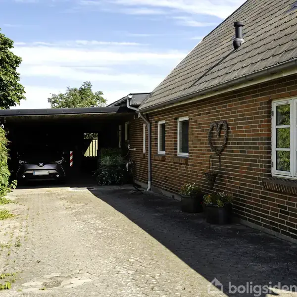 Bil holder parkeret under en carport ved siden af et rødstenshus med flere vinduer. På husets mur hænger en hjerteformet dekoration. To plantekrukker står ved indgangen.