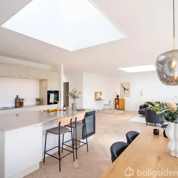 Et moderne køkken-alrum med ø, to barstole og spisebord i forgrunden. En sofa og planter i baggrunden. Lyse elementer og indbygget loftlys skaber en luftig atmosfære.