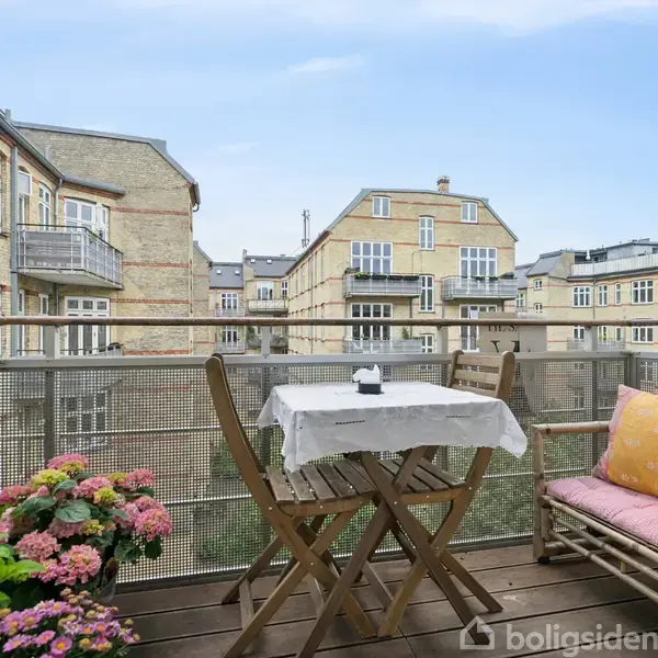 En lys stue med en grå sofa mod en hvid væg dekoreret med plakater. Ved siden af en stol med fåreskind en kurvelignende reol med grøn plante og stearinlys. En rund glastopbord står foran sofaen.