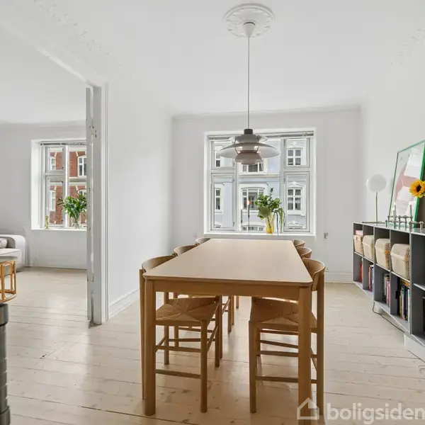 Et bord med stole omkring står i en lys spisestue. Ved siden af er en bogreol med bøger og dekorationer. Store vinduer lader lys ind, og der er friske blomster.