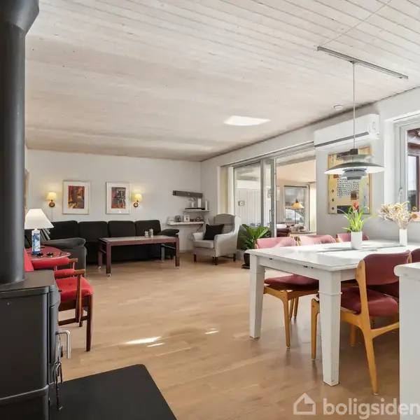 Stue med pejs til venstre, lyserød sofa og sorte lænestole i baggrunden. Spisebord med røde stole står ved store vinduer, der giver udsigt til terrasse.