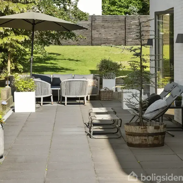 Terrasse med moderne havemøbler under parasol, omgivet af grønne planter i krukker. Stole står på flisebelægning ved siden af husets hvide murvæg, med en velholdt græsplæne i baggrunden.