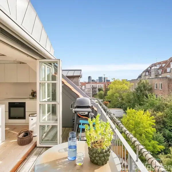 En altan med grill og bord med planter strækker sig ud fra et moderne køkken med åbne dobbeltdøre. I baggrunden ses grønne træer og byhuse under en blå himmel.