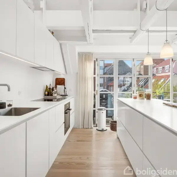 Lyst køkken med hvide skabe og moderne apparater. Store vinduer lukker sollys ind, mens hængelamper oplyser rummet. Køkkenø i centrum, omgivet af træpaneler.