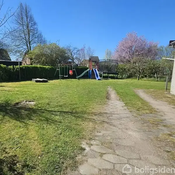 En have med en græsplæne og gyngestativ, omgivet af buske og træer, ved siden af en flisebelagt vej der leder til en bygning.
