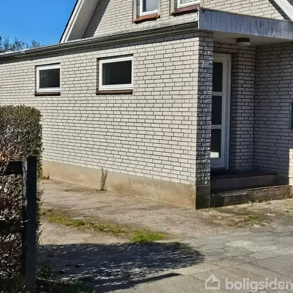 Hus med lyse mursten og to små vinduer, hvor døren står på et indgangsparti med trapper. Omkringliggende gør et fortov med lidt bevoksning op til huset.