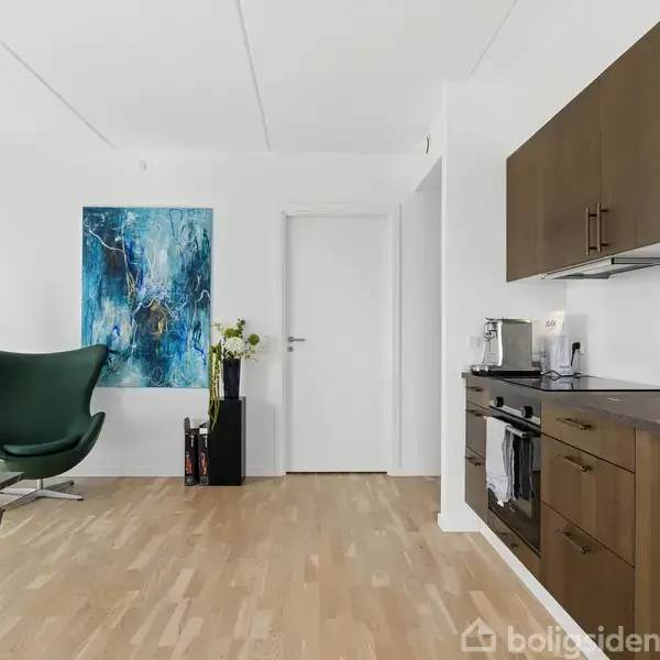 Et moderne køkken med mørke skabe og indbyggede apparater strækker sig langs væggen. Til venstre ses en grøn lænestol ved siden af et blåt maleri på væggen.