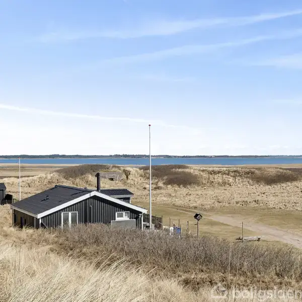 Et sort træhus står stille i klitlandskabet ved en kystlinje, omgivet af sandklitter og med udsigt over havet under en klar, blå himmel.