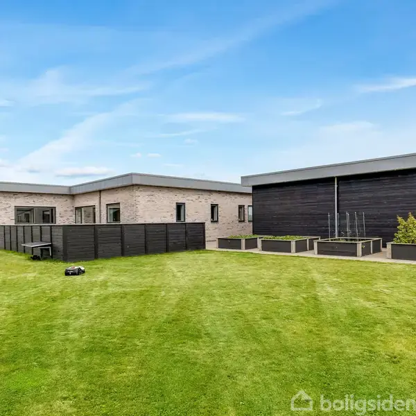 Et moderne hus i lyse mursten står på en velplejet græsplæne. Robotplæneklipper kører foran huset. Til venstre står et sort skur med plantekasser og buske.