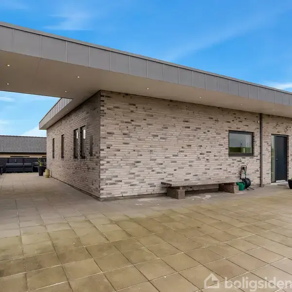 Et moderne enfamiliehus med flade tage og murstensvægge står i en flisebelagt gårdhave. Der er et udendørs vandslangearrangement ved siden af en sort dør, omgivet af blomsterpotter.