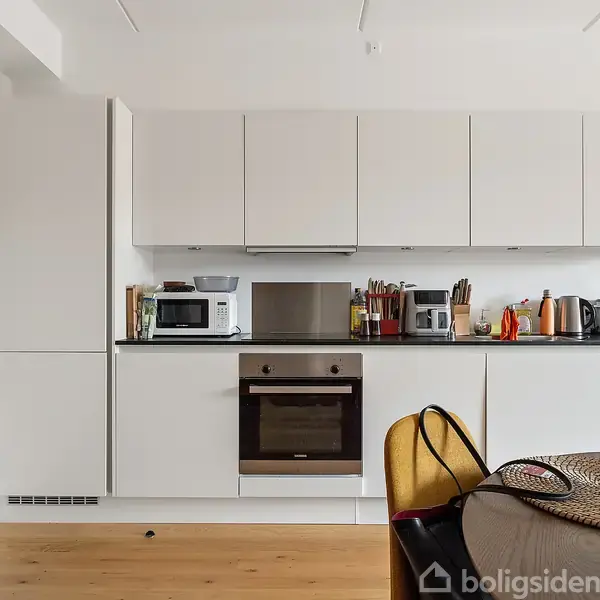 Et moderne køkken med hvide skabe og en integreret ovn; køkkenredskaber og apparater står på bordpladen. En rund spisebord står i forgrunden, omgivet af gyldne stole.