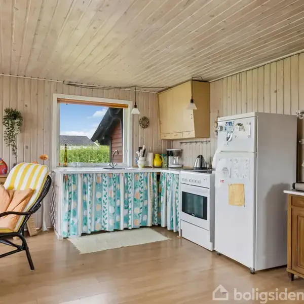Køkken med hvide køkkenelementer og blomstrede gardiner foran vasken. En ovn og køleskab står ved siden af hinanden. En rattan-stol med gul pude står ved siden af et vindue.