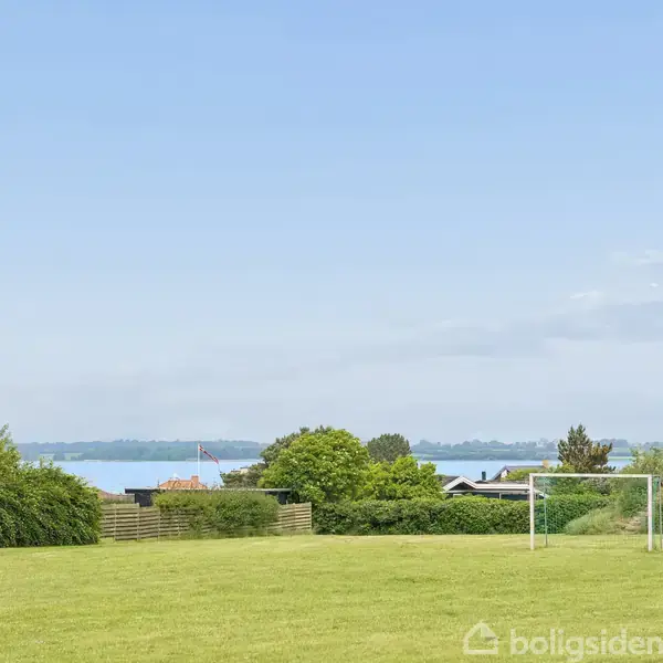 Fodboldmål står på en grøn græsplæne ved et boligområde. I baggrunden ses en udsigt over vand og træer under en blå himmel.