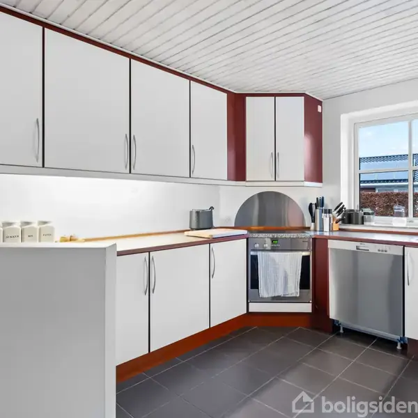 Et moderne køkken med hvide skabe og en integreret ovn. Det har en stor åbning med udsigt til taget af et nabohus. Gulvet er mørkt flisebelagt.