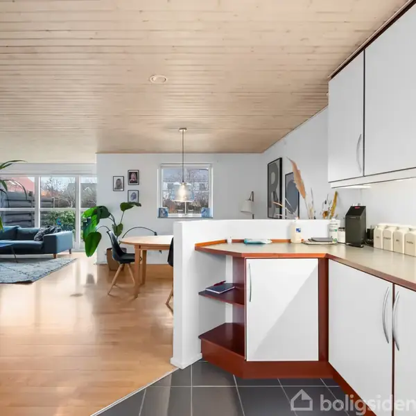 Et moderne køkken i et åbent rum med hvide skabe, der grænser op til en stue med træloft. Der er billeder og magneter på køleskabet og et hjørnebord med kaffemaskine.