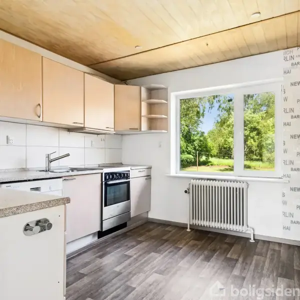 Køkken med træskabe og gasovn står i et rum med lyst trægulv. Et stort vindue viser en grøn have udenfor. Væggene er dekoreret med tekst som "NY," "MILAN," "PARIS."