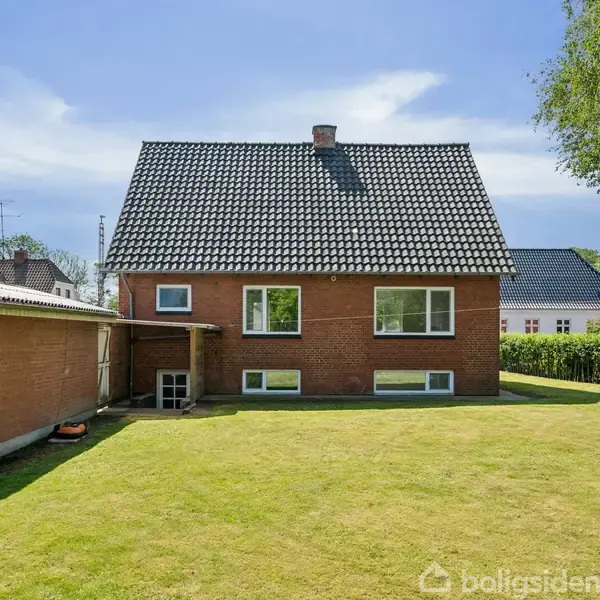 Rødt murstenshus med sort tegltag står stille i en velplejet have med grønt græs, flankeret af et udhus til venstre og træer til højre under en klar blå himmel.